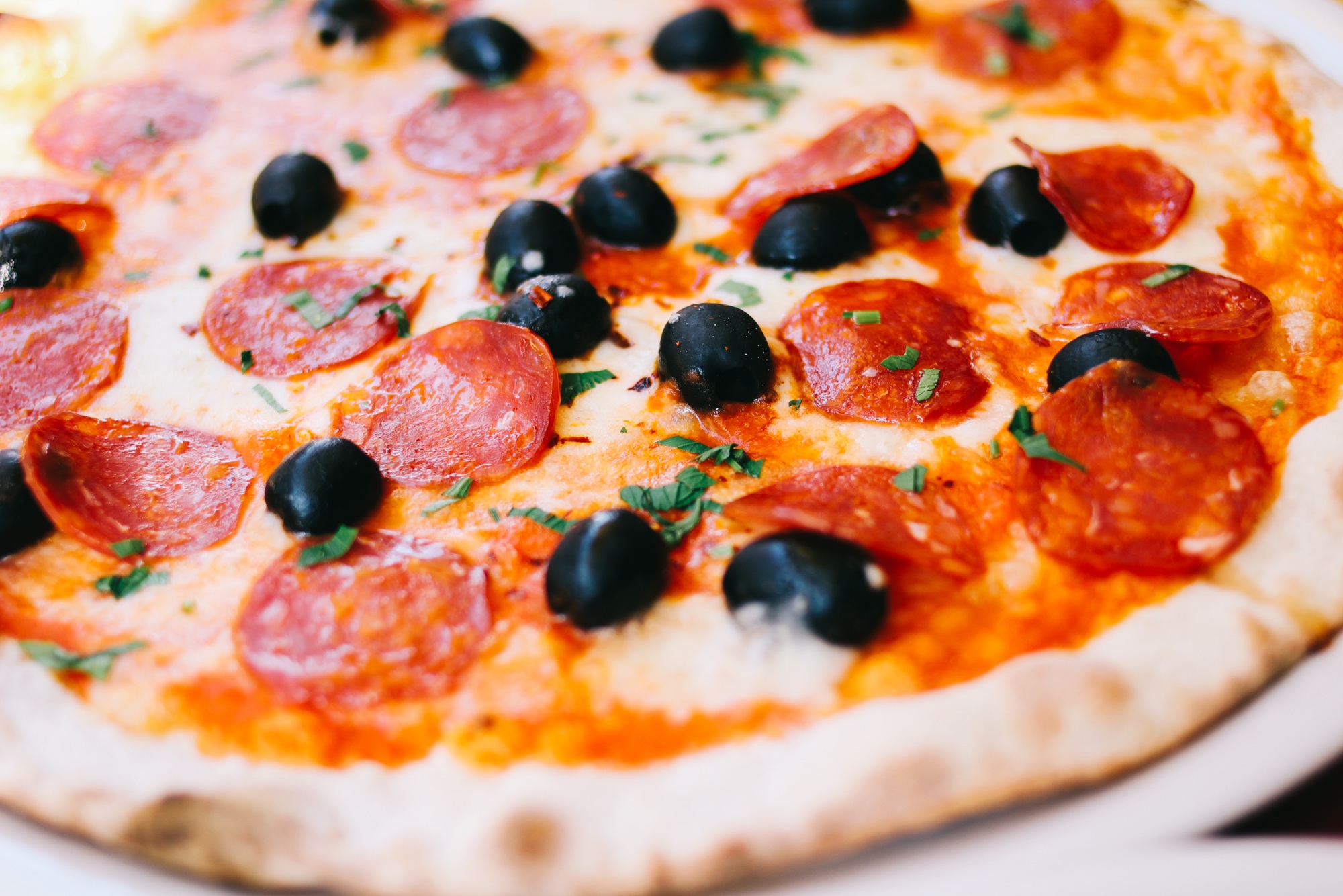 Foto aproximada de uma pizza. Sua base é de molho vermelho e queijo mussarela, coberta por pepperoni e salpicada com azeitonas pretas e manjericão