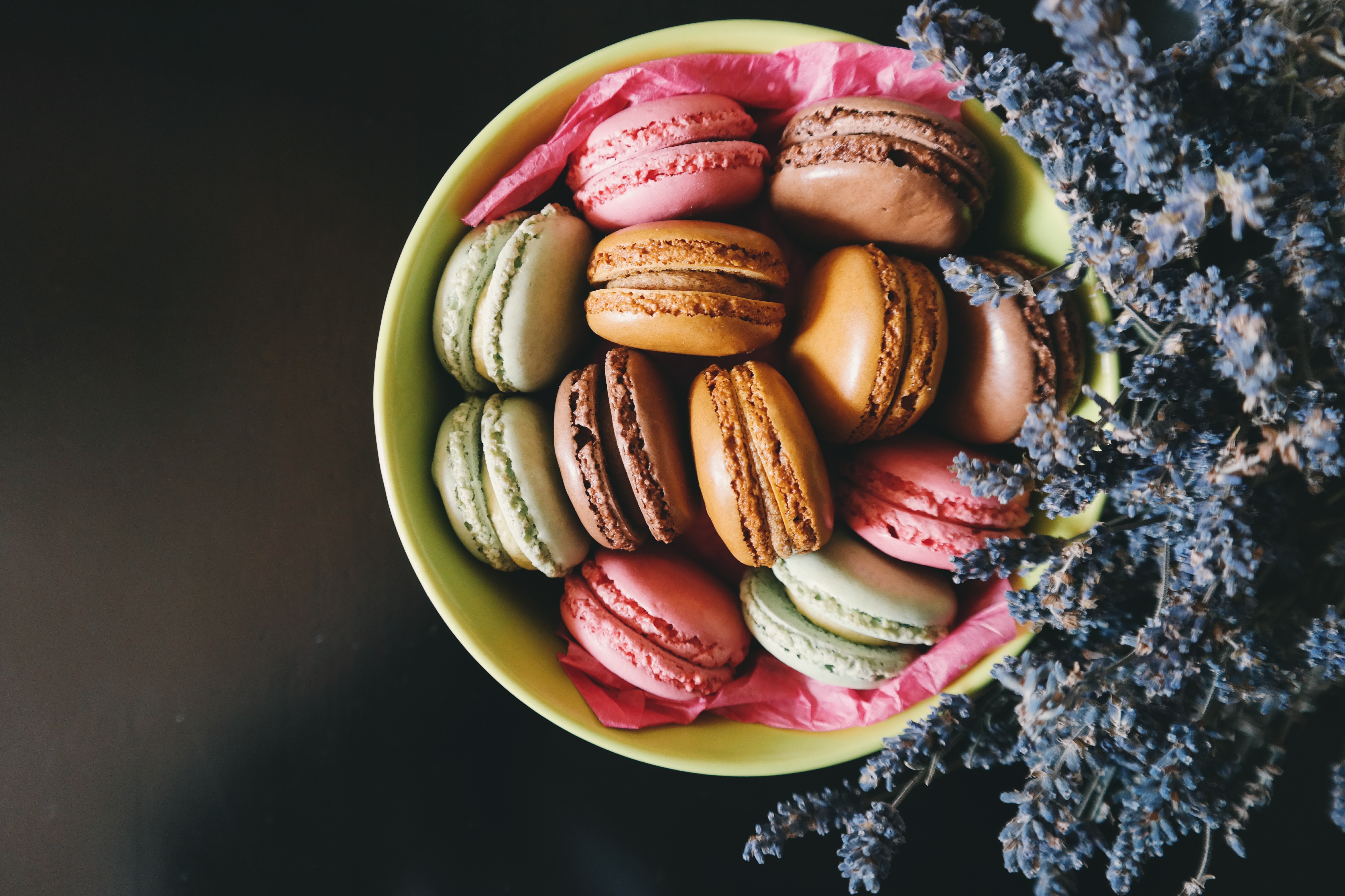 Foto de um bowl cheio de macarrons visto de cima. Dentro do bowl existem quatro sabores de macarrons: morango, na cor rosa; limão, na cor verde; chocolate, na cor marrom; caramelo, na cor bege
