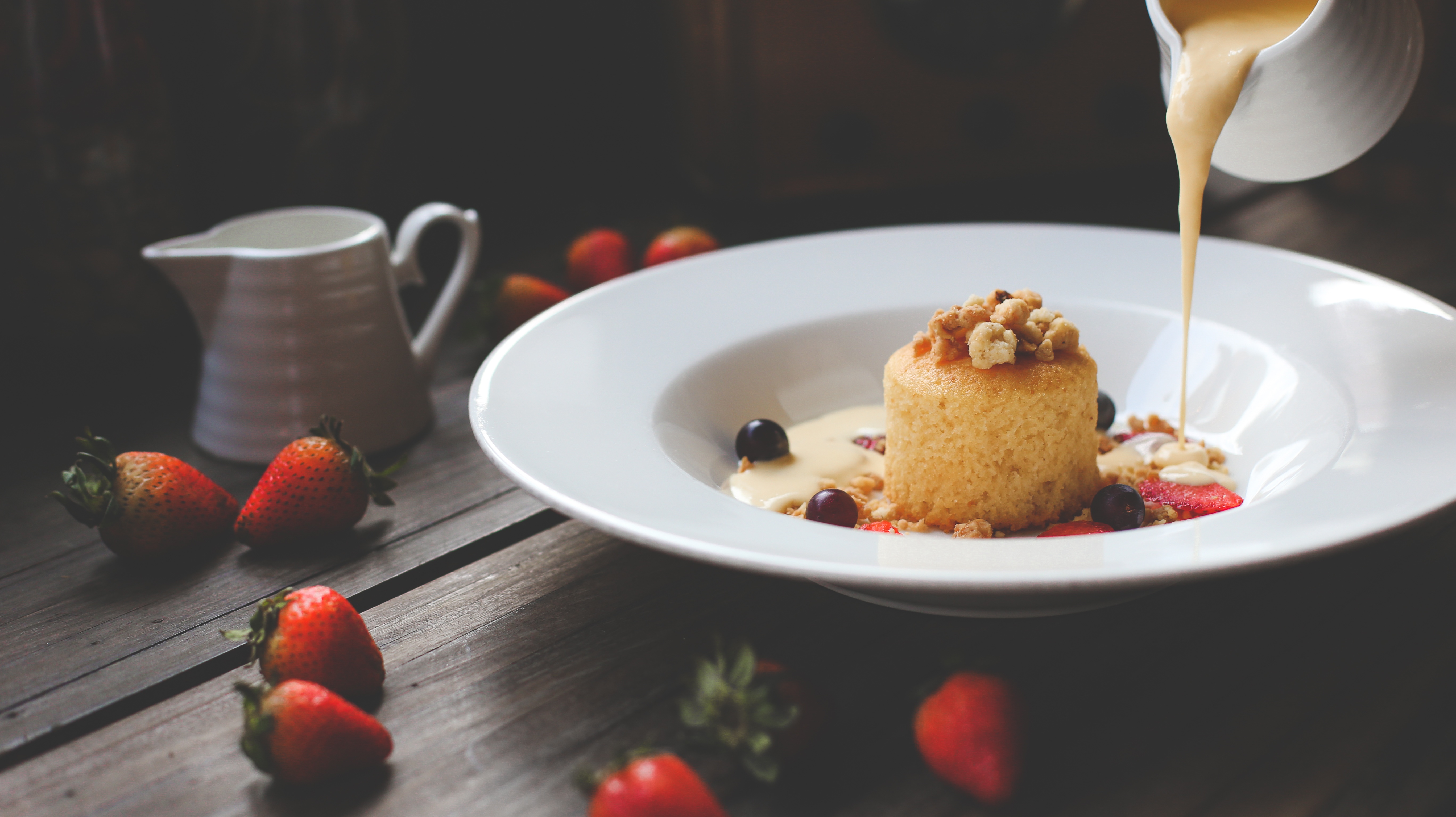 Foto de um prato de petit gateau claro com frutas vermelhas. Ao fundo é possível ver uma jarra despejando calda clara sobre o petit gateau. Em cima da mesa também há morangos e outra jarra de calda