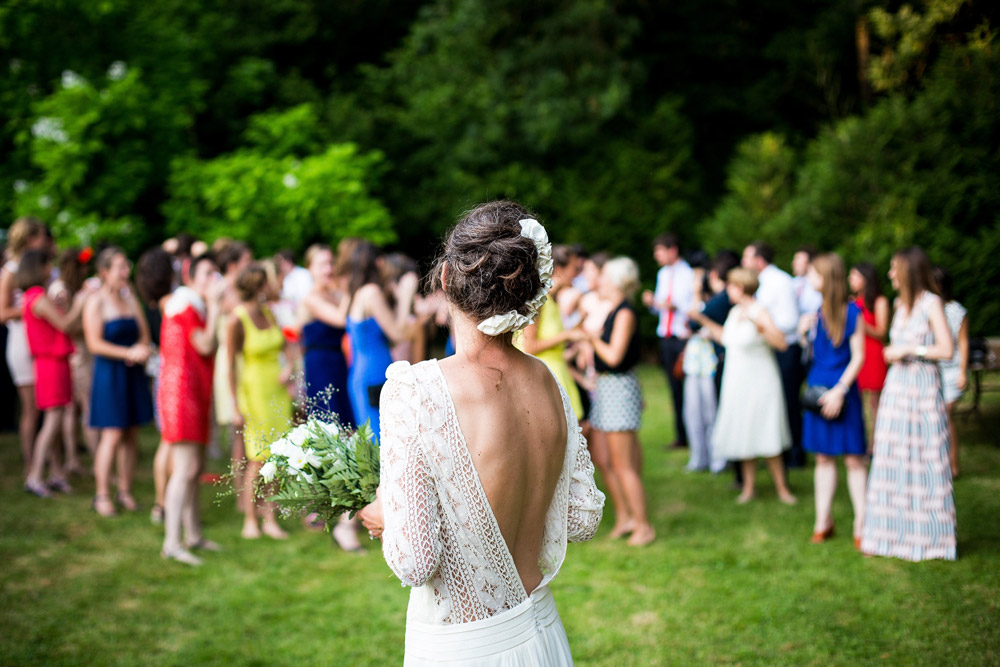 Foto de um casamento ao ar livre. O foco principal é a noiva com um buquet. Os convidados aparecem desfocados no fundo