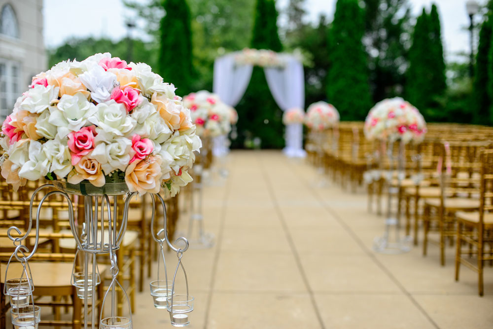 Foto da decoração de um casamento ao ar livre. O foco principal é um arranjo de flores nas cores branco, rosa e salmão. Ao fundo, desfocado, estão as arvores, o altar e as cadeiras para os convidades